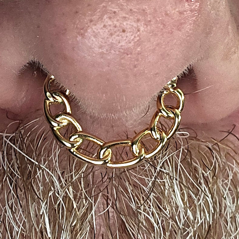 A close-up of a persons nose and mustache, featuring a large gold chain-link septum ring piercing through the nose.