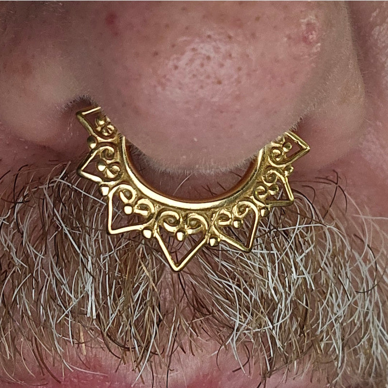 Close-up of a persons face showing a gold plated, ornate tribal septum clicker in a man's septum piercing.