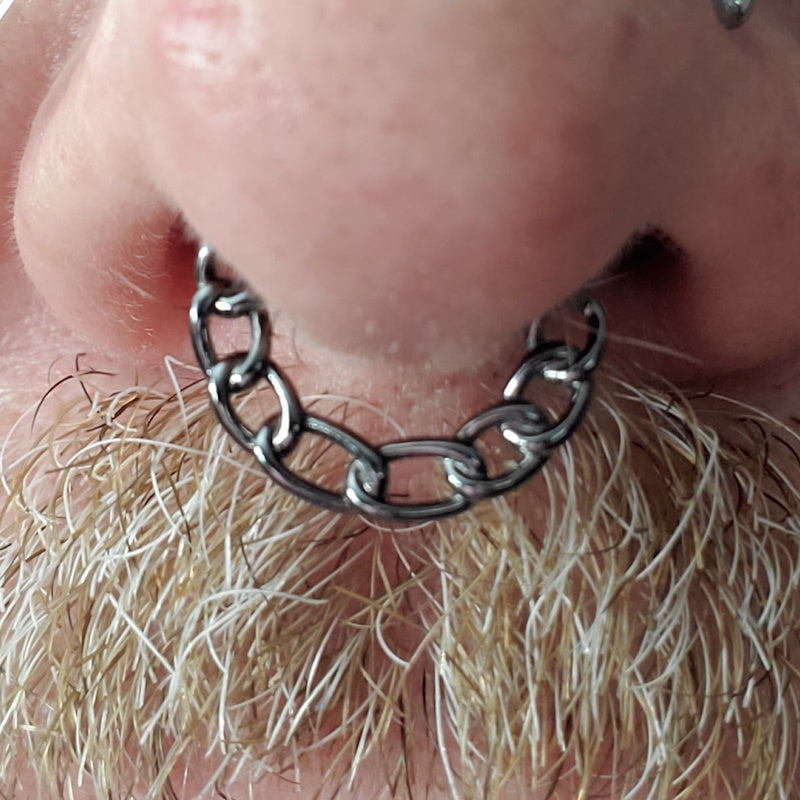 Close-up of a persons nose with a large chain-link ring septum piercing, above a thick, light-colored mustache.