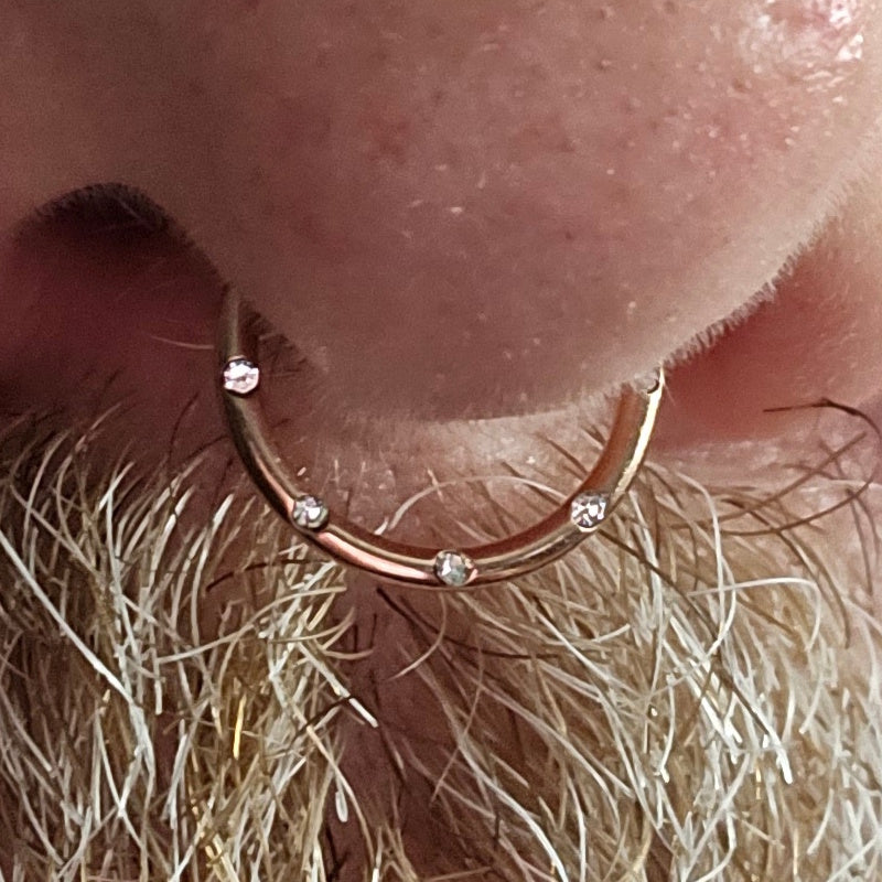 A close-up of a septum nose piercing with a rose gold hoop ring, adorned with small clear gemstones. Blonde facial hair is visible below the nose.