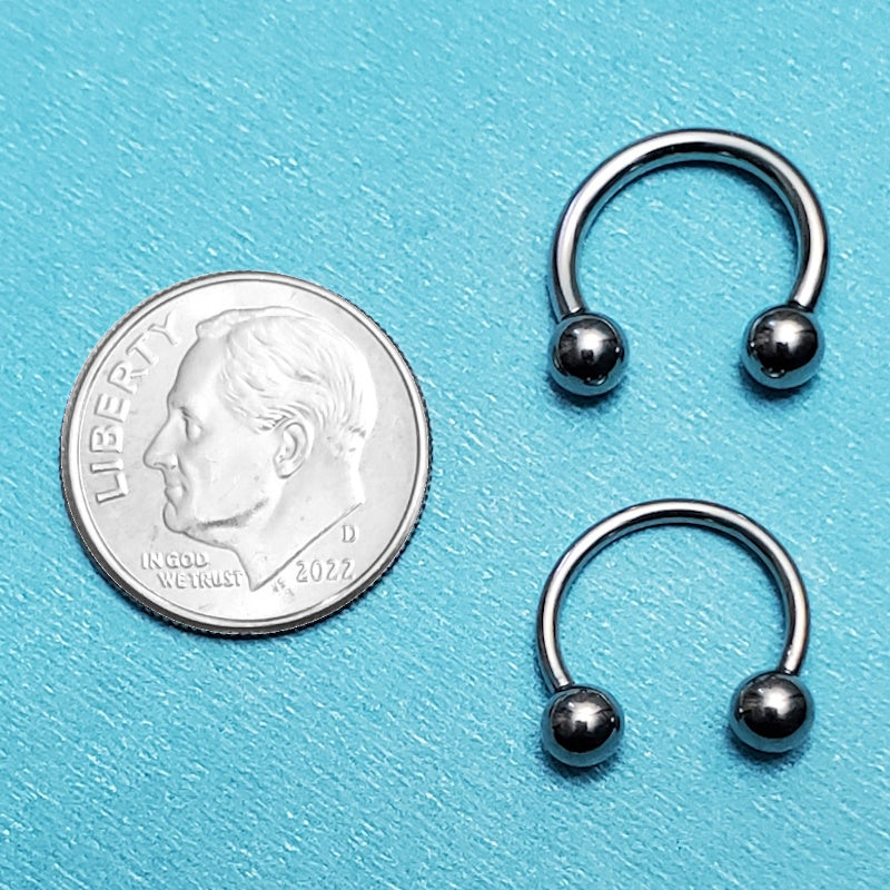 Two sizes of a surgical steel septum ring pictured on an aqua background next to a dime for a size comparison.