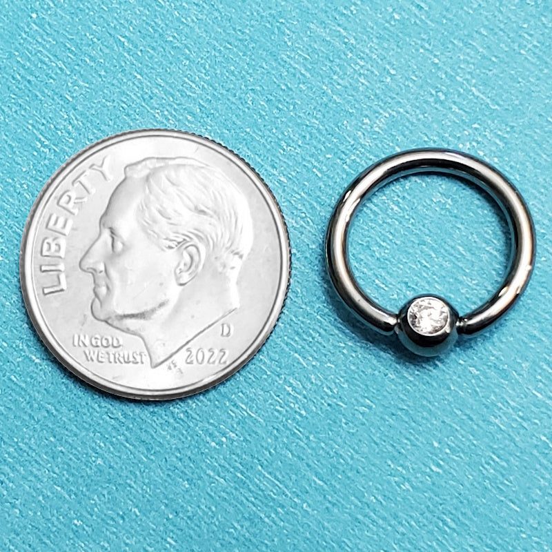 A 14 gauge septum ring pictured on an aqua background next to a dime for a size comparison.