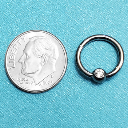 A 14 gauge septum ring pictured on an aqua background next to a dime for a size comparison.