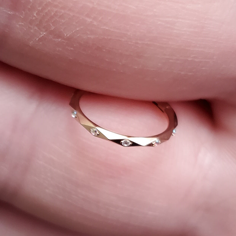 A titanium 16g rose gold septum ring featuring cubic zirconia accented diamond faceted cuts pictured between the fingers of a hand.