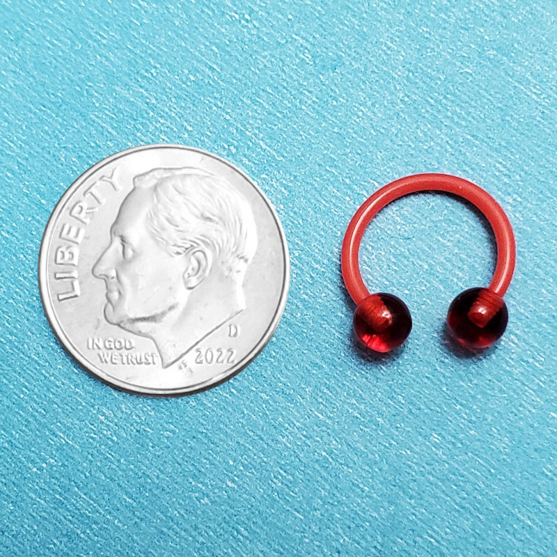 An acrylic red septum ring pictured on an aqua background next to a dime for a size comparison.