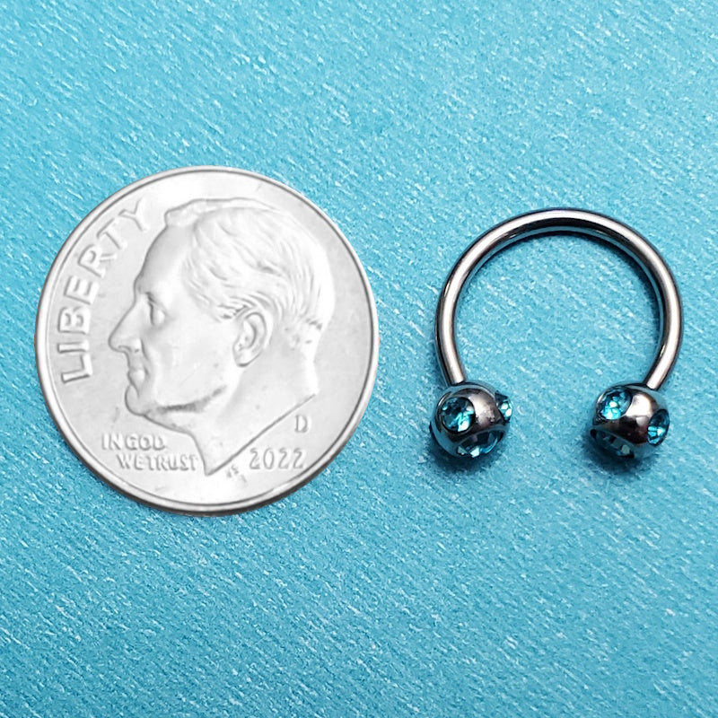 An aqua gem septum ring pictured on an aqua background next to a dime for a size comparison.