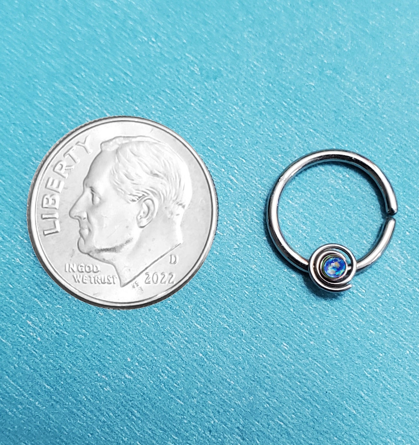 A bendable blue opal septum ring pictured on an aqua background next to a dime for size comparison.