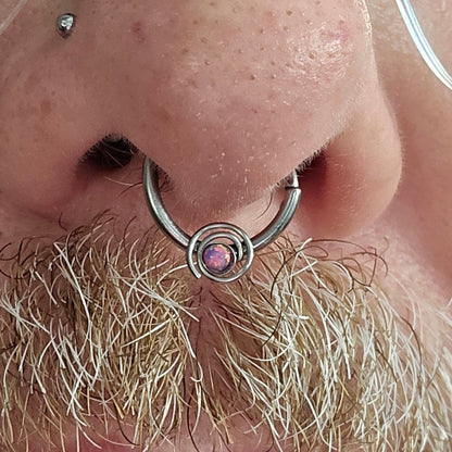 A close-up of a nose with a silver septum ring featuring a pink synthetic opal in the center. The person has a light moustache with mixed blonde and white hairs and a silver nostril piercing.