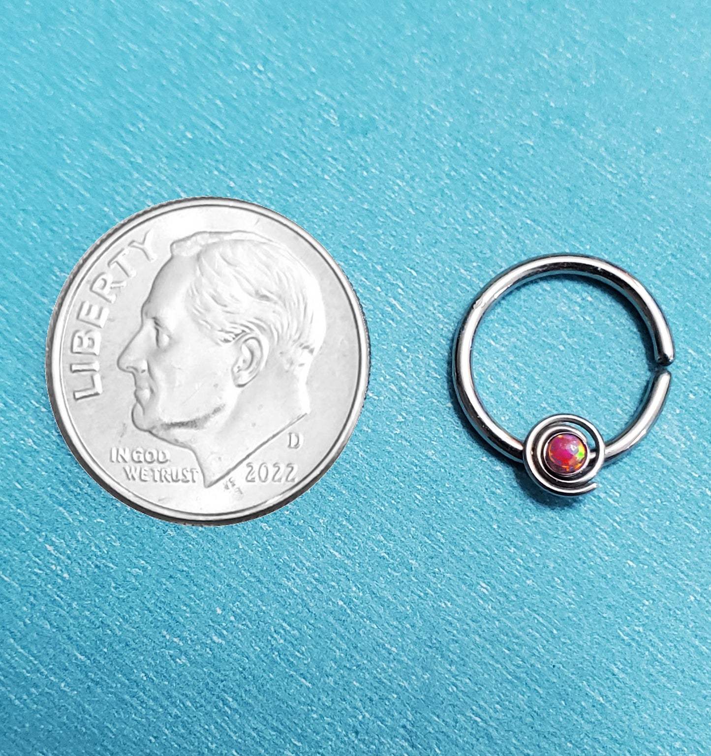 A bendable opal septum ring pictured on an aqua background next to a dime for a size comparison.