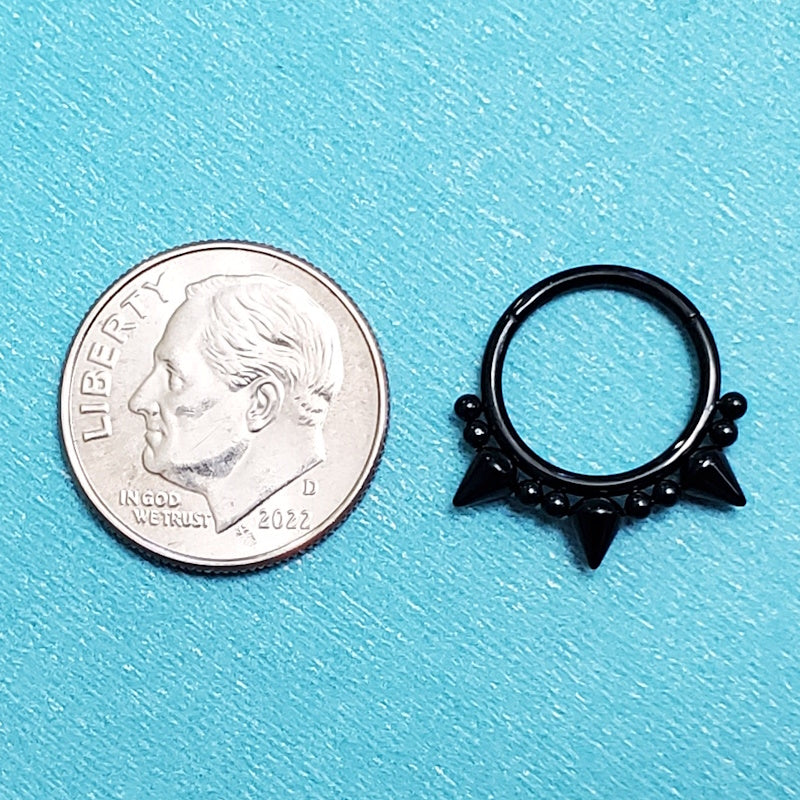 A black spike septum ring pictured on an aqua background next to a dime for a size comparison.