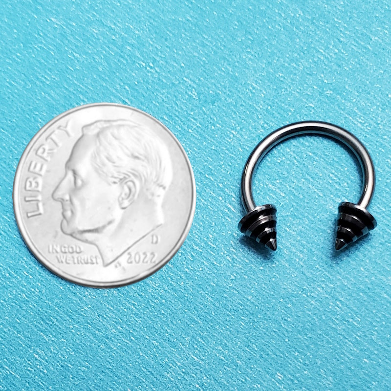 A black stripe spike septum horseshoe pictured on an aqua background next to a dime for a size comparison.