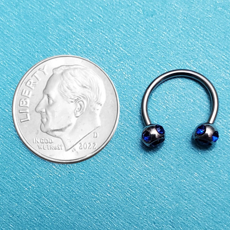 A blue gem septum ring pictured on an aqua background next to a dime for a size comparison.