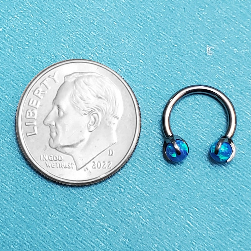 A blue opal septum ring pictured on an aqua background next to a dime for a size comparison.