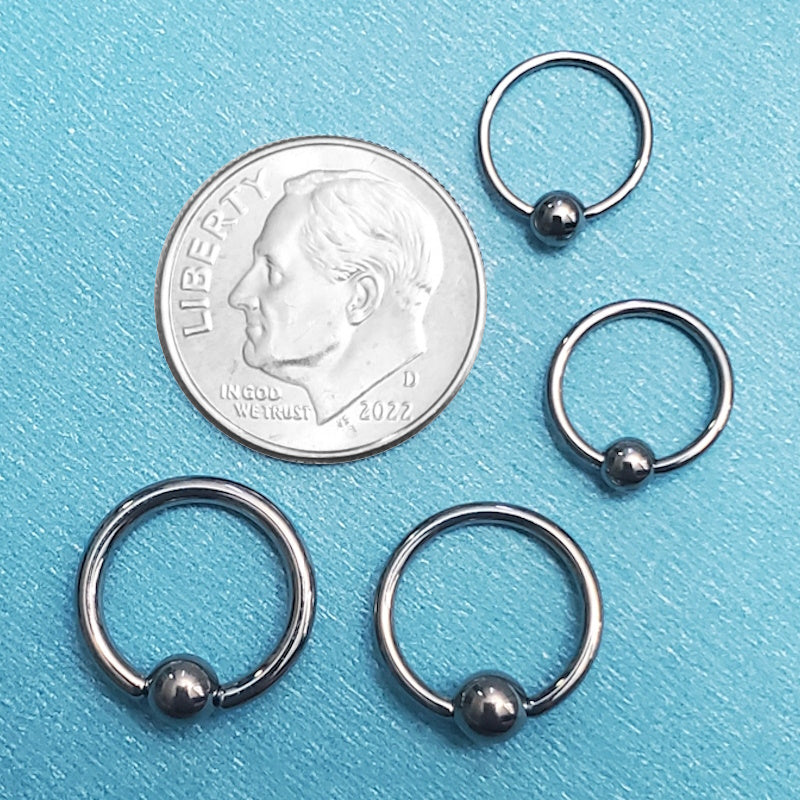 Five sizes of captive bead titanium segment rings pictured on an aqua background next to a dime for a size comparison.