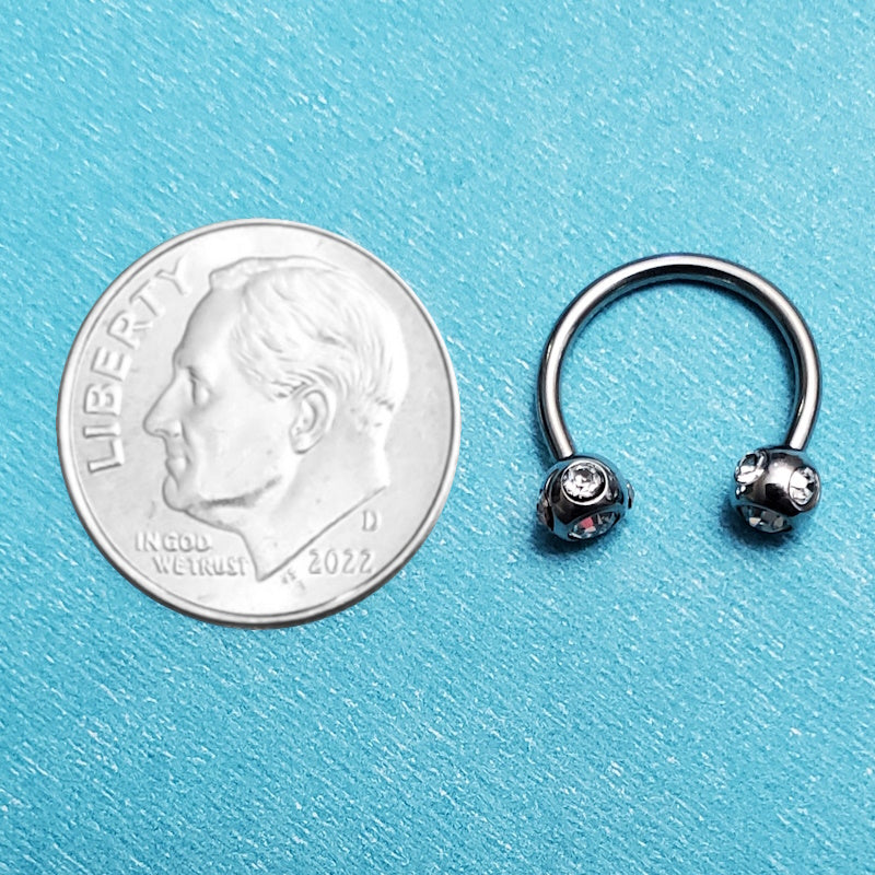 A clear gem septum ring pictured on an aqua background next to a dime for a size comparison.