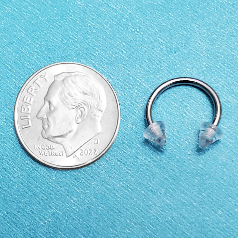 A clear spike septum ring pictured on an aqua background next to a dime for a size comparison.