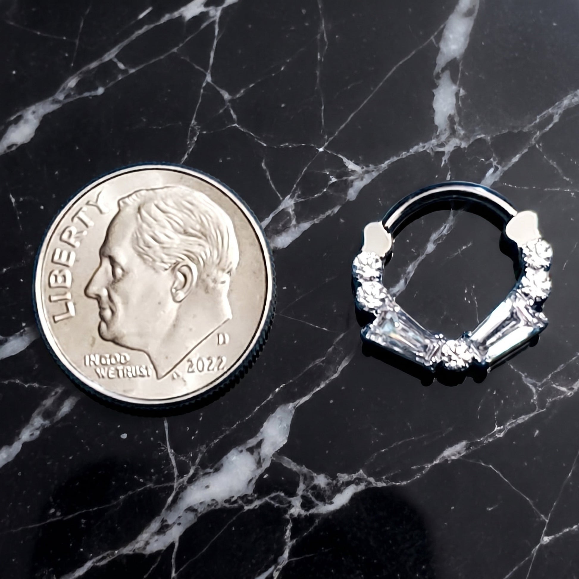 A cubic zirconia septum ring with baguette and round cuts pictured next to a dime for size comparison on a black marble countertop.