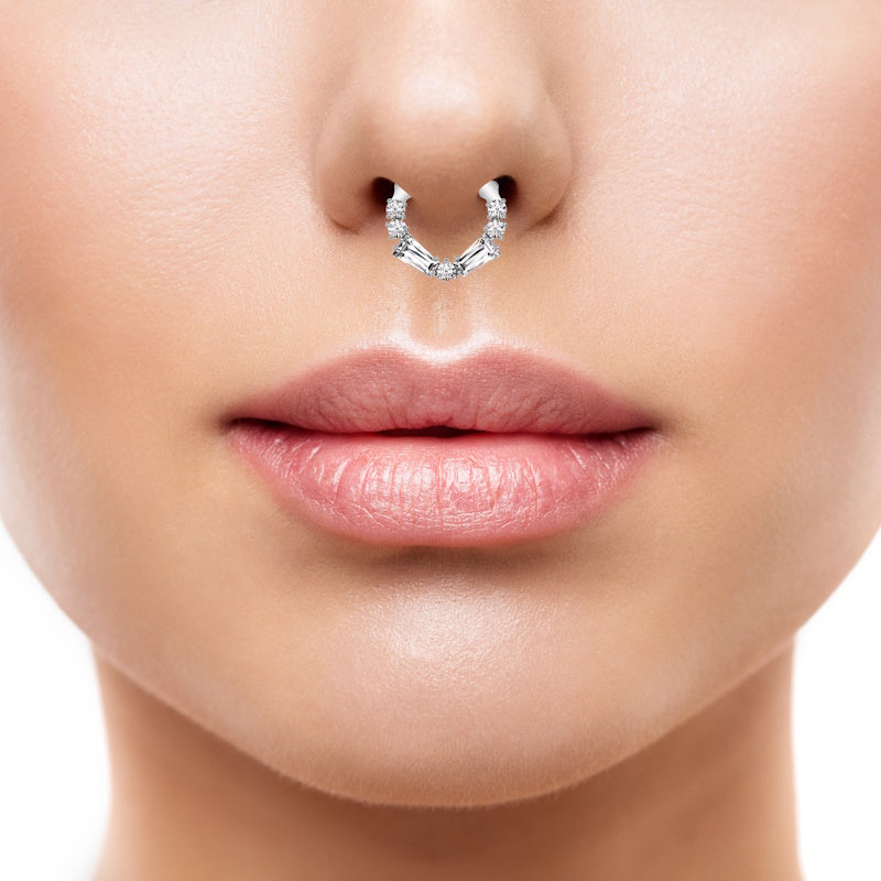 A beautiful cubic zirconia septum ring with round cubic zirconia and 2 baguette cubic zirconia on the bottom half of a septum hoop pictured in the septum piercing of a female.