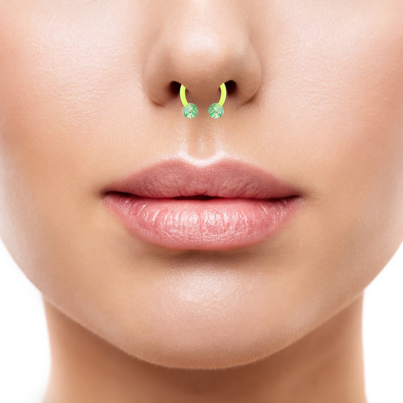 A glitter green septum ring with a horseshoe shaped design and a ball on each end filled with glitter pictured in the septum piercing of a female.