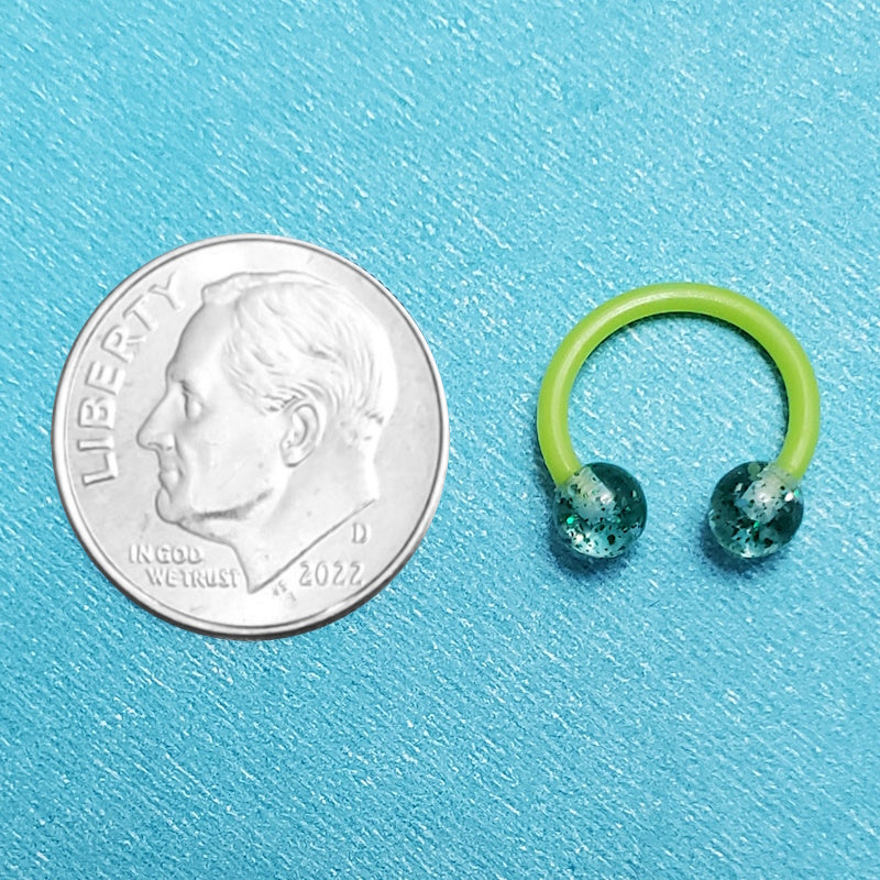 A glitter green septum ring pictured on an aqua background next to a dime for a size comparison.