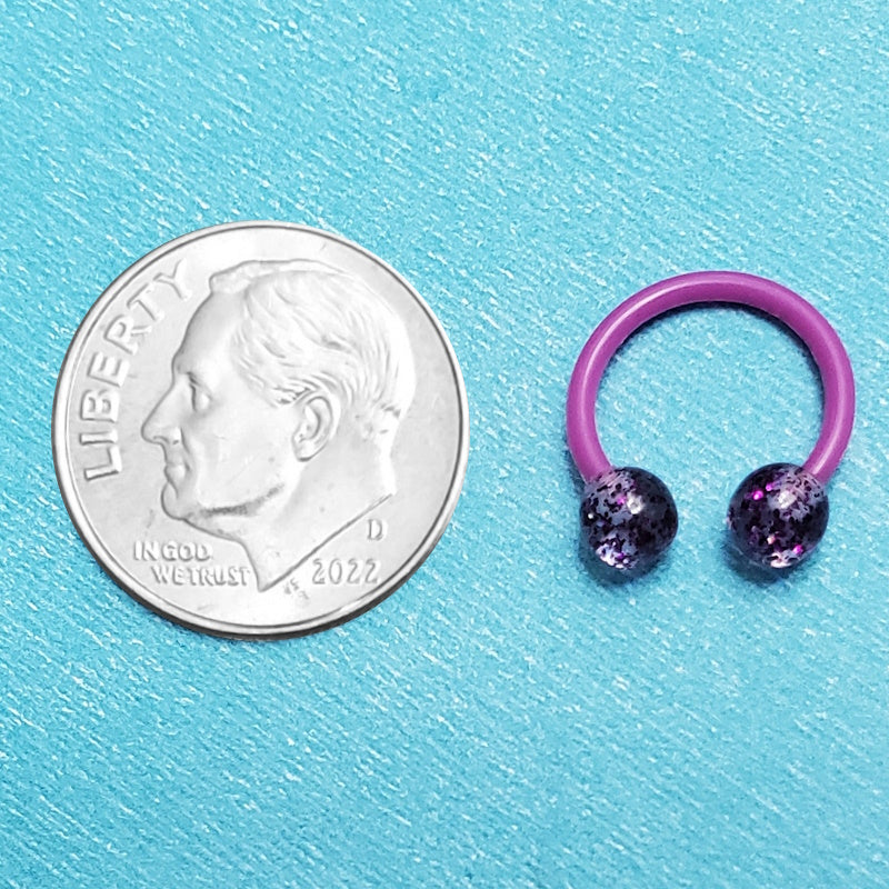 A glitter purple septum ring pictured on an aqua background next to a dime for a size comparison.