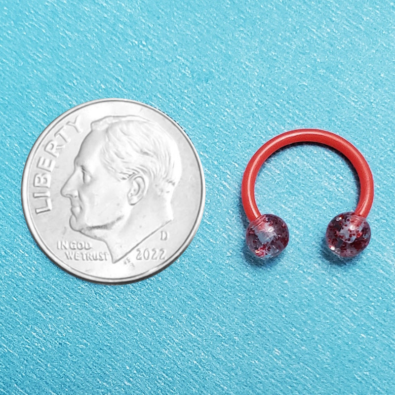 A glitter red septum ring pictured on an aqua background next to a dime for a size comparison.