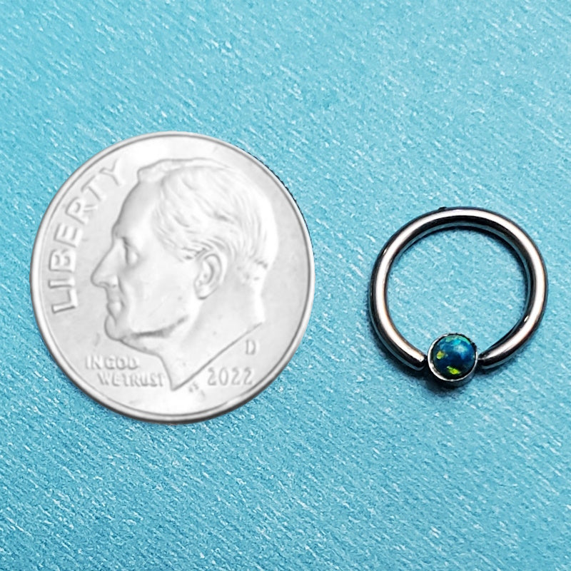 A green opal septum ring pictured on an aqua background next to a dime for a size comparison.