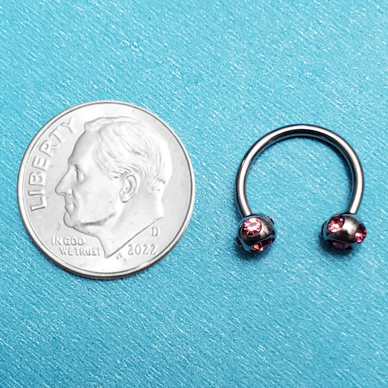 A pink gem septum ring pictured on an aqua background next to a dime for a size comparison.