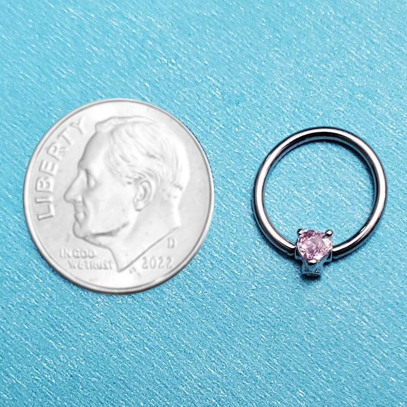 A pink heart septum ring pictured on an aqua background next to a dime for a size comparison.