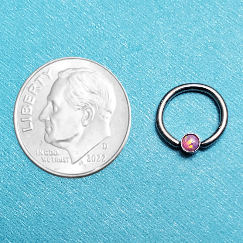 A pink opal septum ring pictured on an aqua background next to a dime for a size comparison.