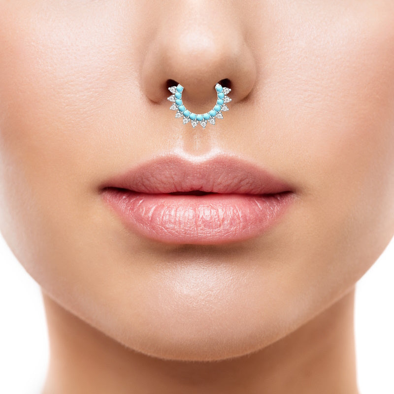 A platinum turquoise septum ring featuring multiple turquoise stones and cubic zirconia lined around the bottom of the bendable hoop pictured in the septum piercing of a woman.