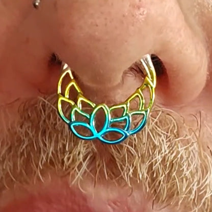 A close-up of a persons nose with a colorful metallic septum ring shaped like a flower. The person has facial hair, a nose stud, and wears glasses.