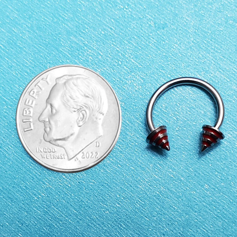 A red stripe spike septum horseshoe pictured on an aqua background next to a dime for a size comparison.