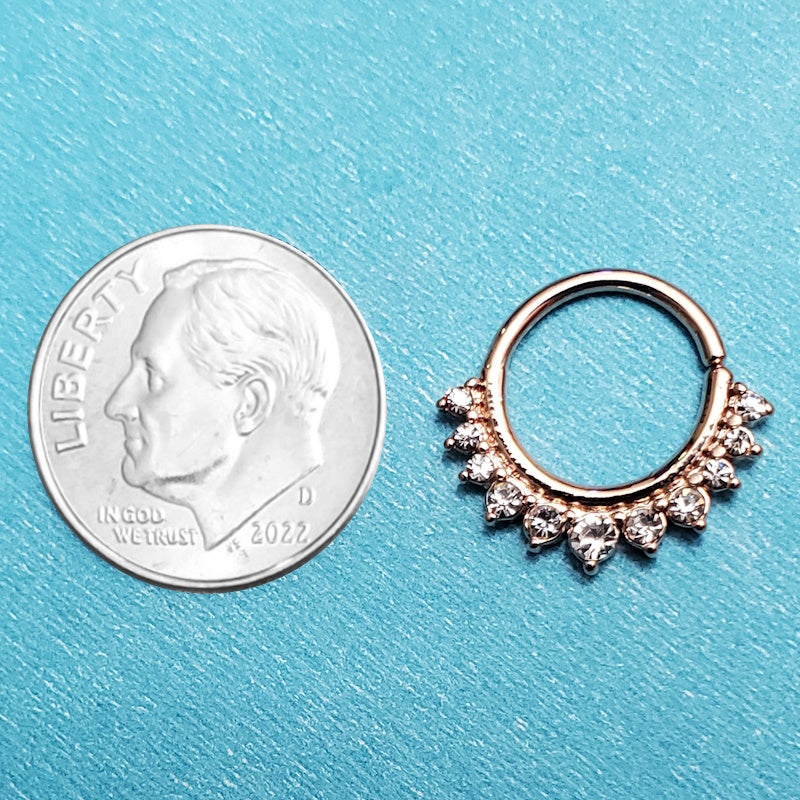 A rose gold crystal septum ring pictured on an aqua background next to a dime for a size comparison.