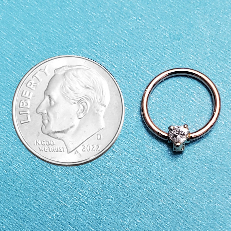 A rose gold cz heart septum ring pictured on an aqua background next to a dime for a size comparison.