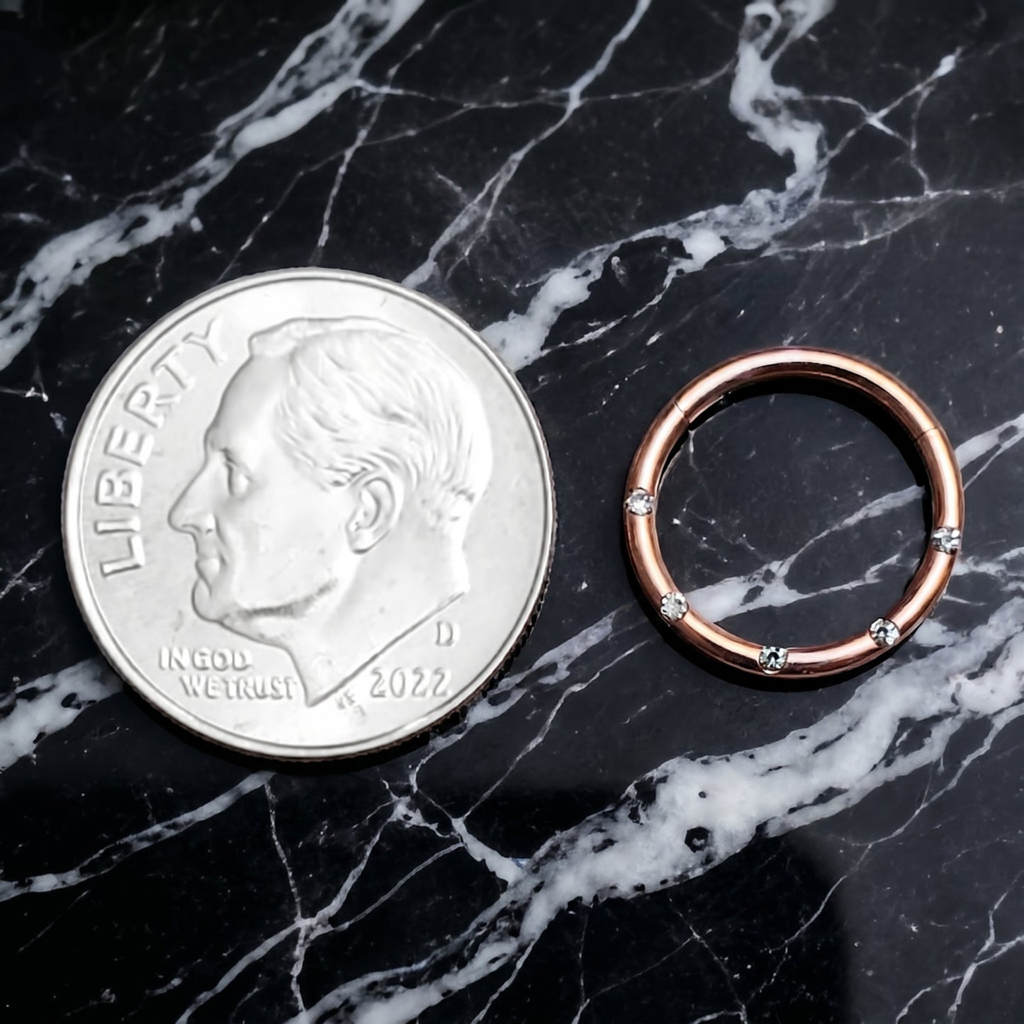 Rose Gold Hinged Segment Ring Cubic Zirconia 16G 316L Surgical Steel placed on a black marble countertop next to a US dime for size comparison.