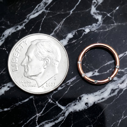 Rose Gold Hinged Segment Ring Cubic Zirconia 16G 316L Surgical Steel placed on a black marble countertop next to a US dime for size comparison.