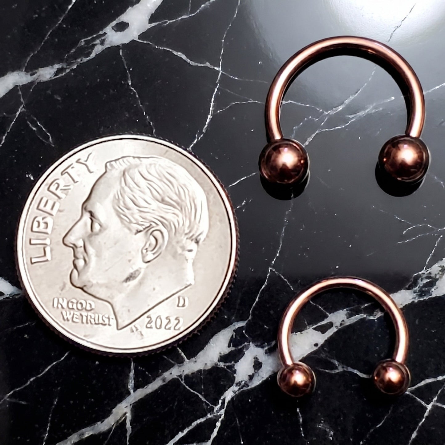 Two Rose Gold Horseshoe Circular Barbell Septum Rings pictured on a black marble background next to a dime for size comparison.