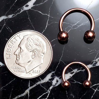 Two Rose Gold Horseshoe Circular Barbell Septum Rings pictured on a black marble background next to a dime for size comparison.