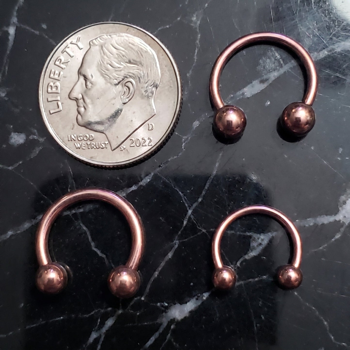Three Rose Gold Horseshoe Septum Rings with Ion Plating Over 316L Surgical Steel, pictured on a black marble background with a dime for a size comparison.