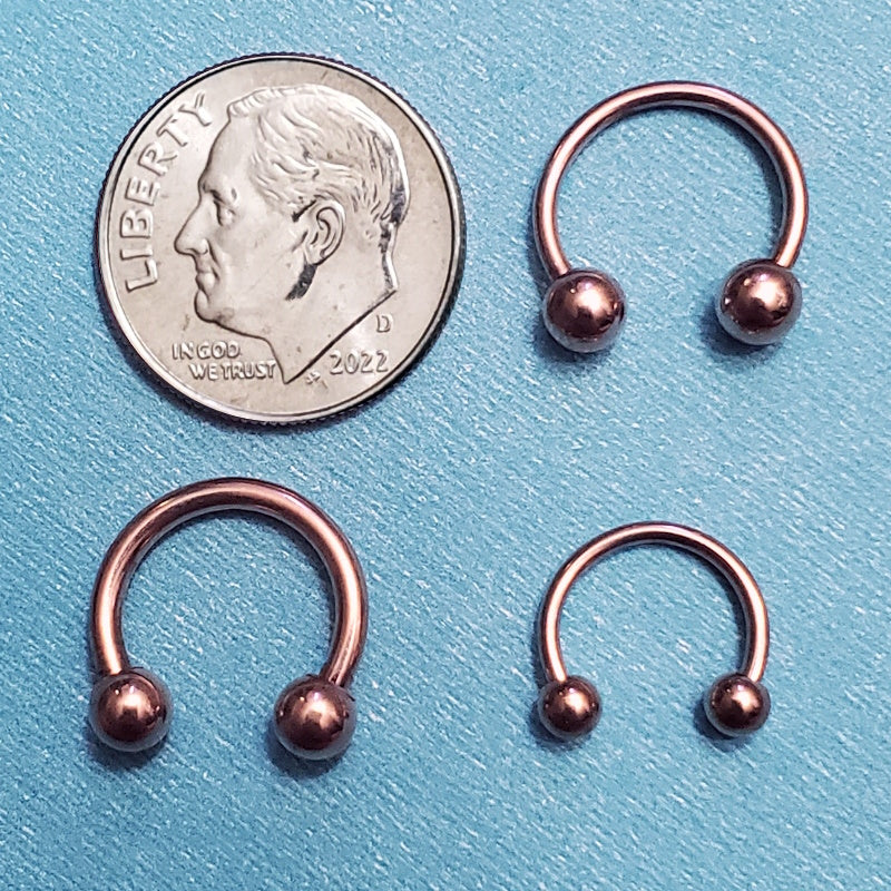 Three sizes of a rose gold horseshoe septum rings pictured on an aqua background next to a dime for a size comparison.