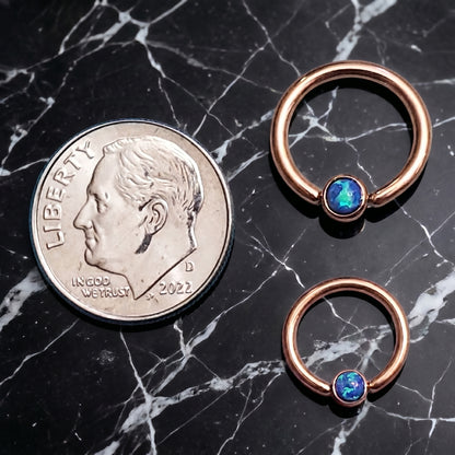 Two sizes of a rose gold opal captive bead septum ring next to a dime for size comparison on a black marble countertop.