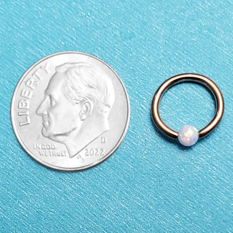 A rose gold opal captive septum ring pictured on an aqua background next to a dime for a size comparison.