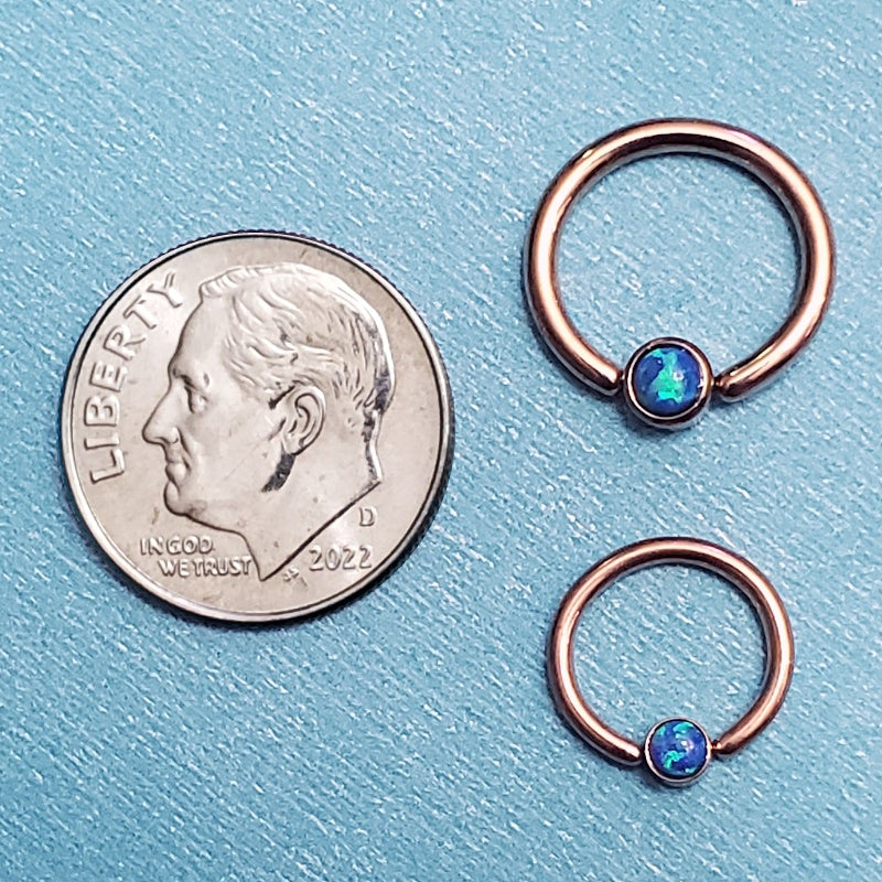 Two sizes of a rose gold opal septum rings pictured on an aqua background next to a dime for a size comparison.