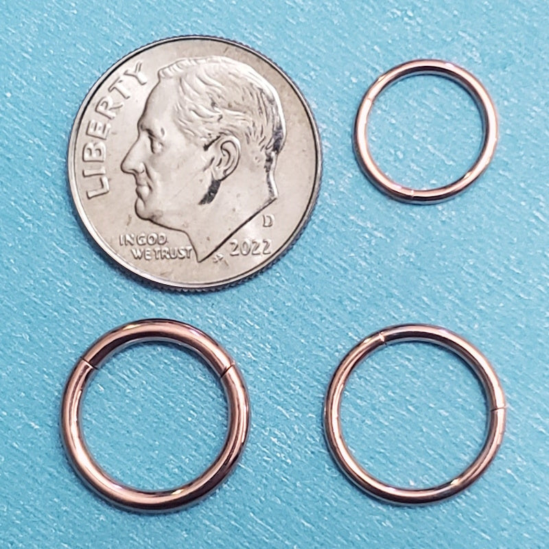 Three sizes of a rose gold septum hoops pictured on an aqua background next to a dime for a size comparison.