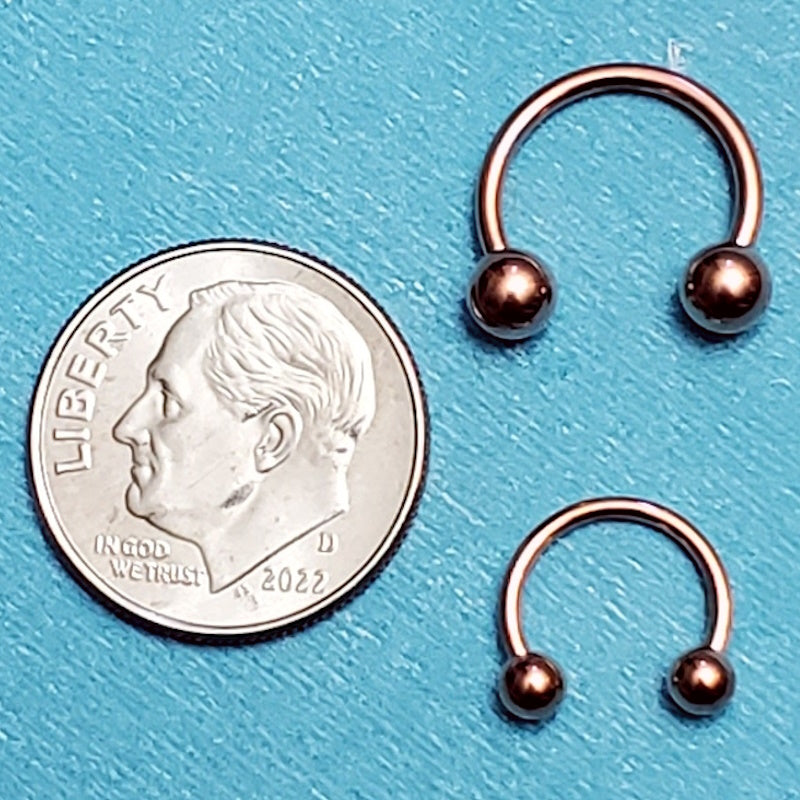 Two sizes of a rose gold septum horseshoes pictured on an aqua background next to a dime for a size comparison.