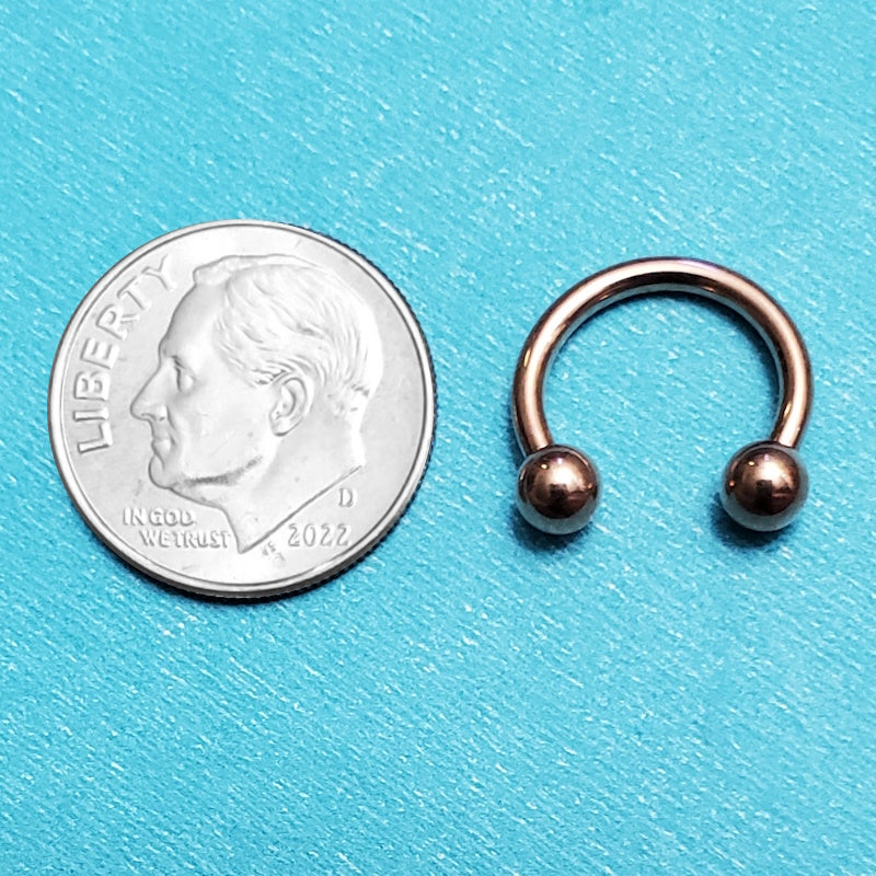 A rose gold septum ring pictured on an aqua background next to a dime for a size comparison.