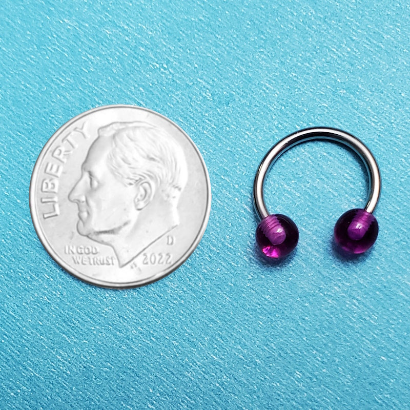 A septum piercing circular barbell pictured on an aqua background next to a dime for a size comparison.