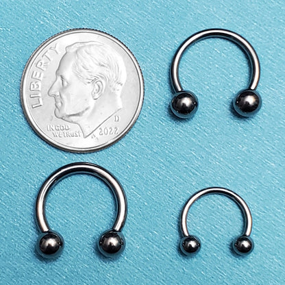 Three sizes of stainless steel septum rings pictured on an aqua background next to a dime for a size comparison.