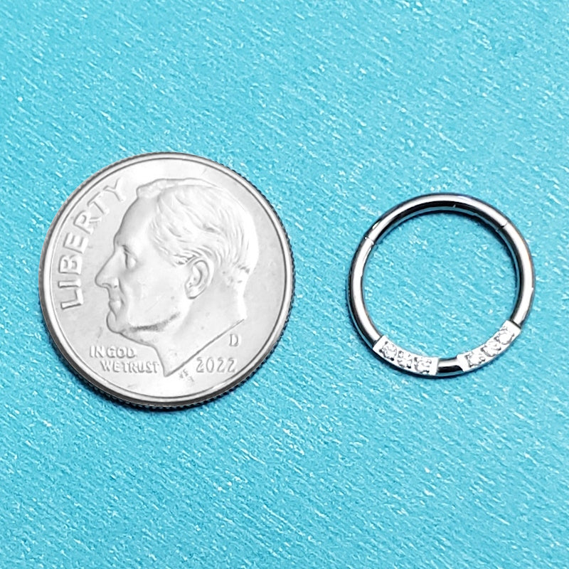 A surgical steel 16g segment ring pictured on an aqua background next to a dime for a size comparison.